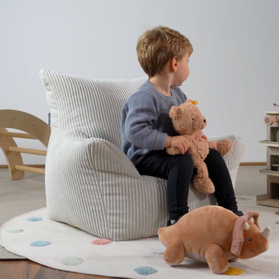 Pouf enfant en velours côtelé beige dans une chambre d'enfant. Un enfant assis sur le pouf, regardant loin de la caméra et tenant un ours en peluche marron à la main